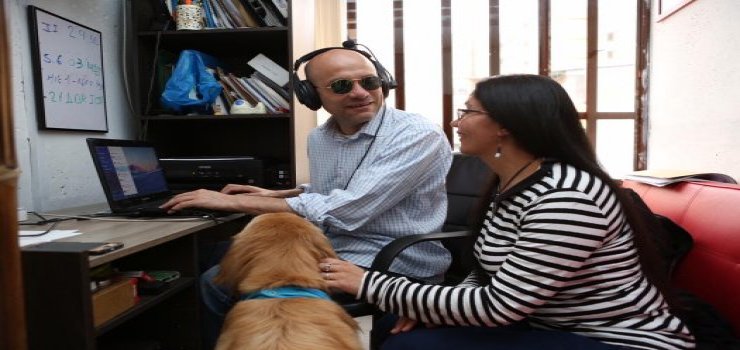 Hombre sentado frente a escritorio con computador, con su esposa y perro guía